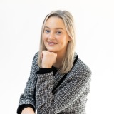 Professional headshot of Sarah, smiling confidently in a checkered blazer, photographed in a bright studio setting.