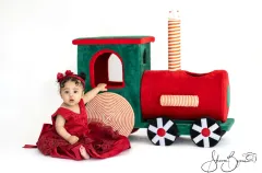 Smiling toddler girl in red lace dress sitting beside a festive Christmas train during a studio photoshoot