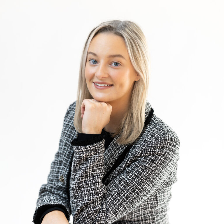 Professional headshot of Sarah, smiling confidently in a checkered blazer, photographed in a bright studio setting.