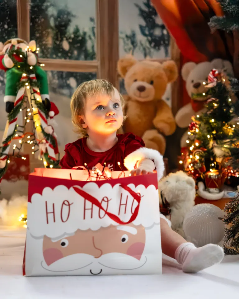 Toddler sitting with a glowing Santa gift bag in a magical Christmas studio scene.