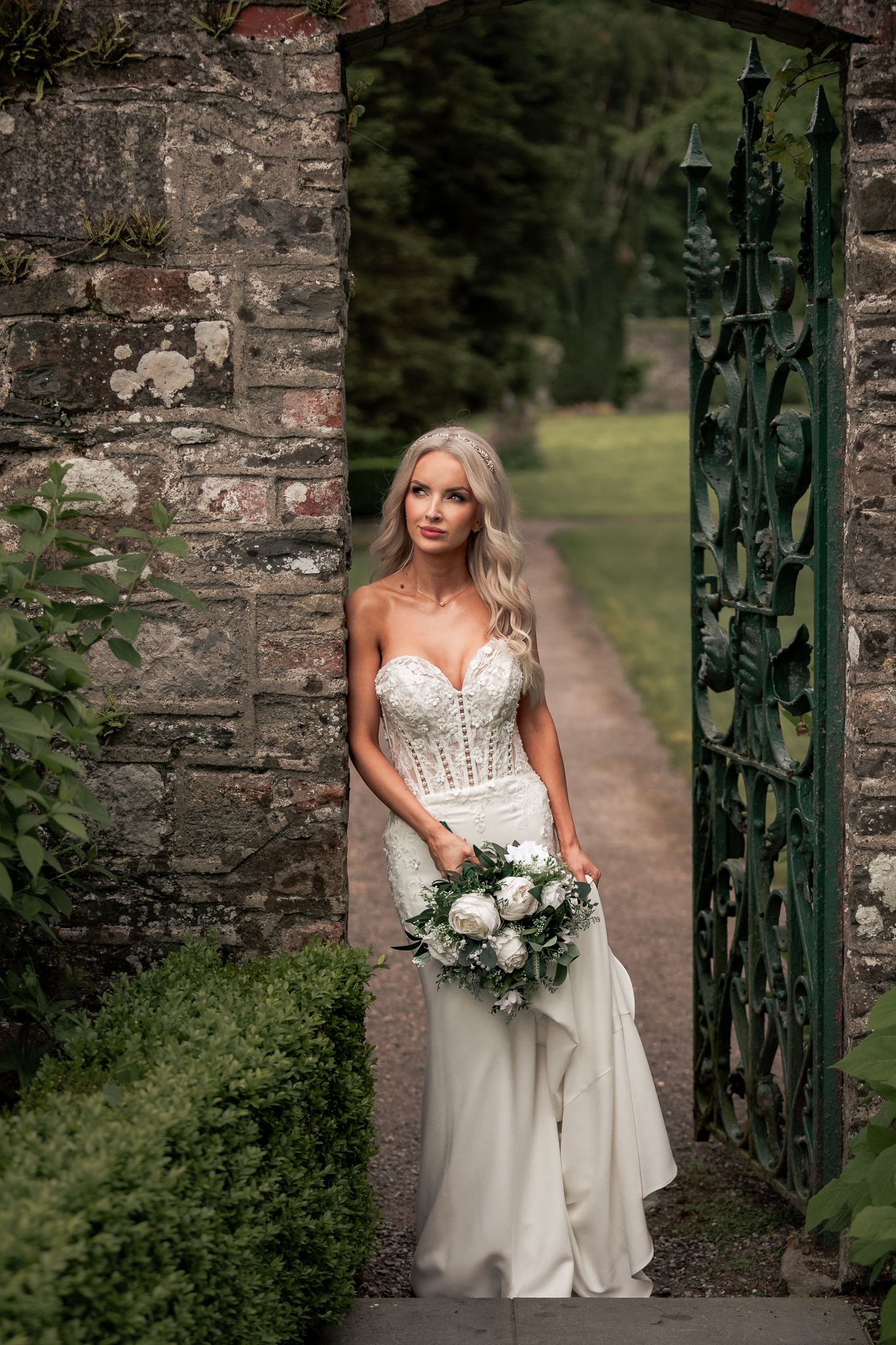 Bride in a fitted lace gown with a bouquet beside an ornate iron gate in a stone wall