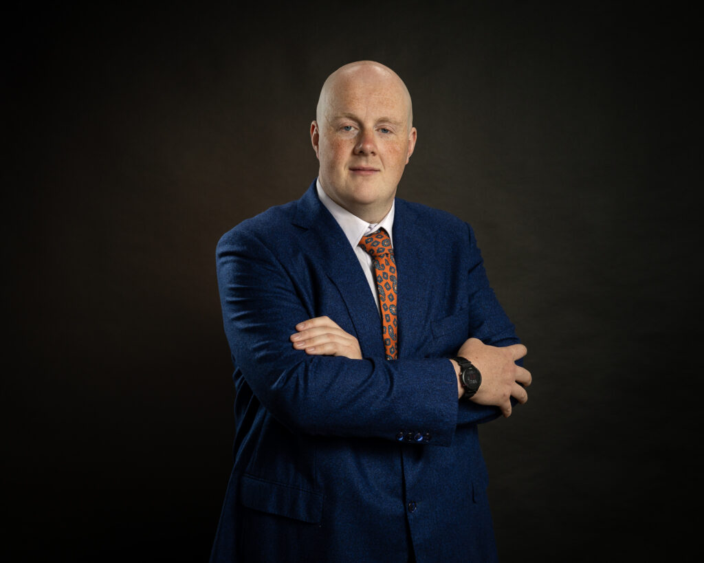 Confident professional headshot of Ross with arms crossed, navy suit, studio near Dublin