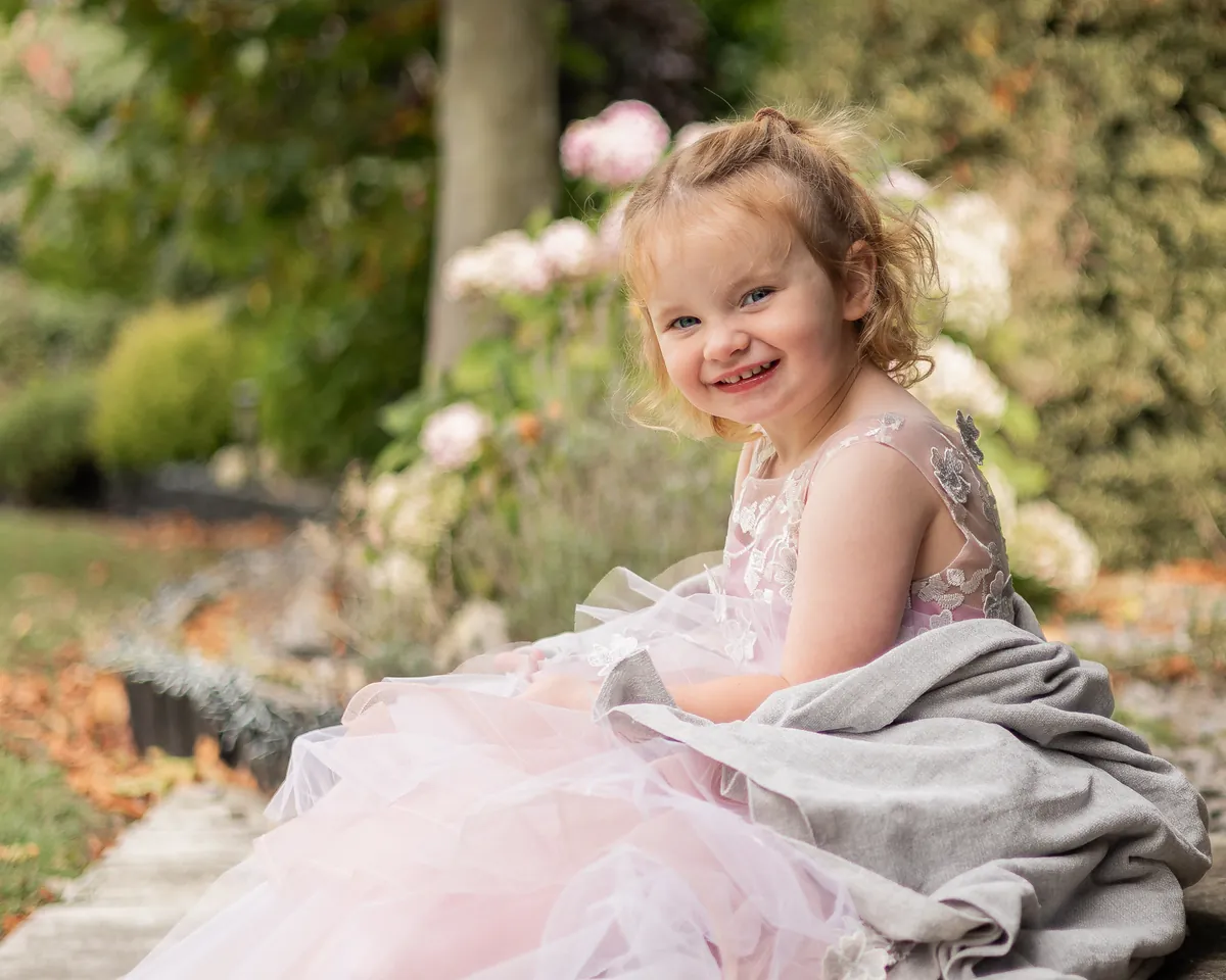 Toddler in a pale pink tulle gown sitting on a garden path and laughing over her shoulder