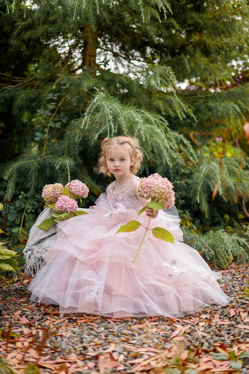 Two year old girl in a pale pink tulle gown sitting under a conifer holding a hydrangea, with fallen autumn leaves around her