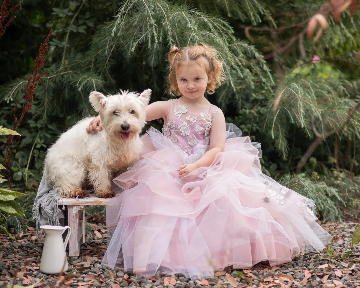 Toddler in a pale pink gown sitting beside a West Highland terrier on a garden bench