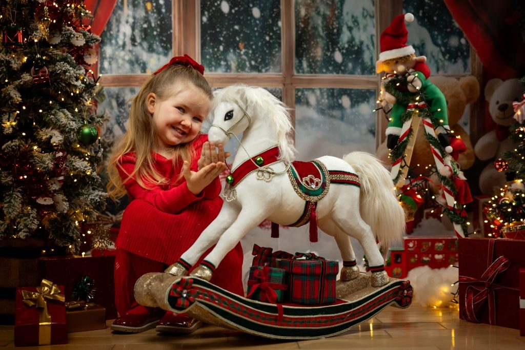Young girl laughing beside a white rocking horse in a Christmas studio set