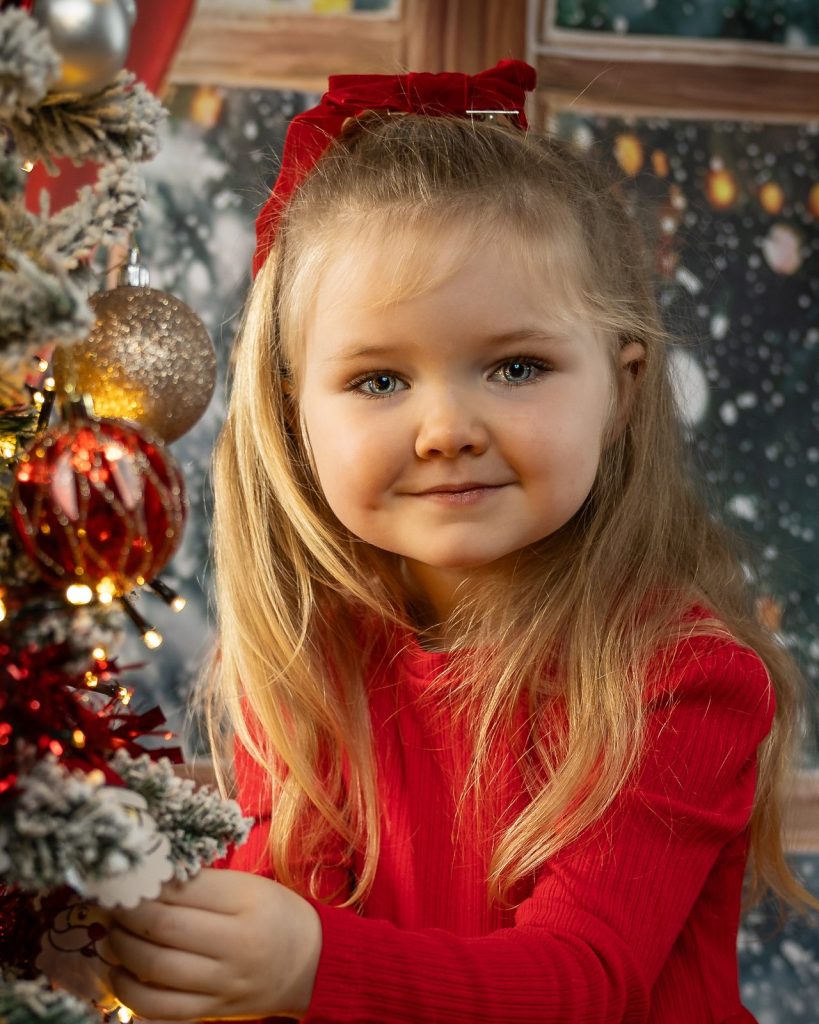 Young girl with a red velvet bow smiling beside a lit Christmas tree