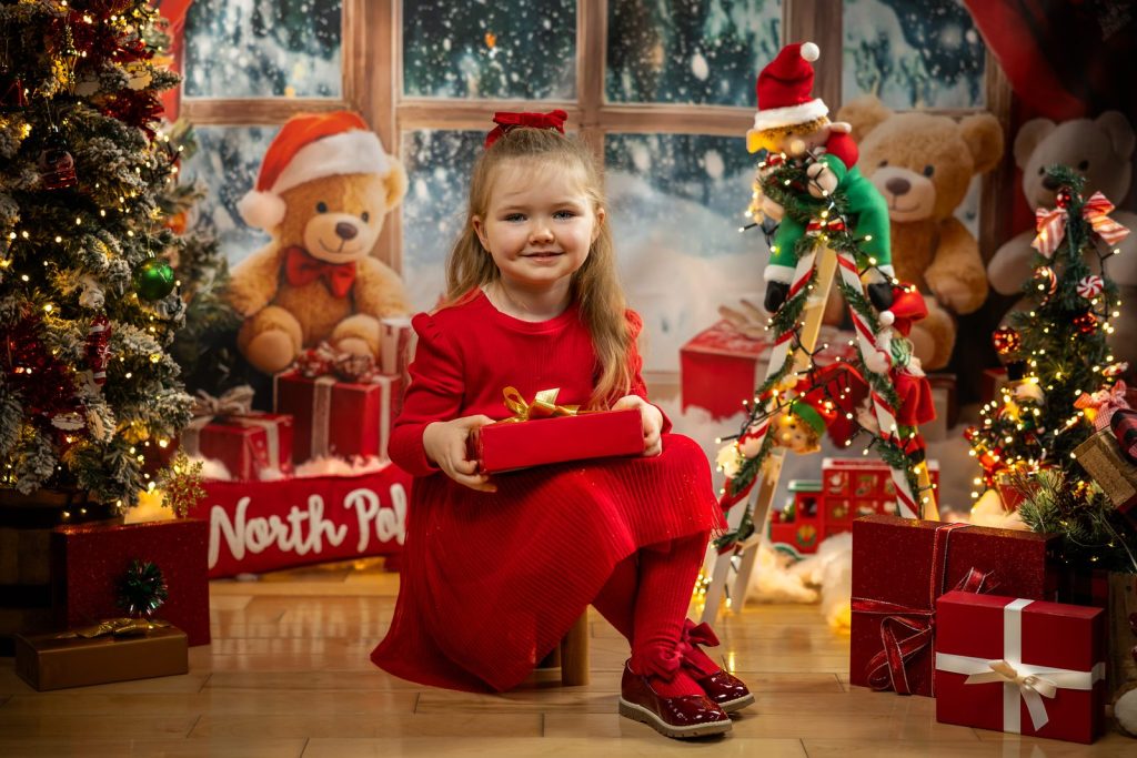 Girl in a red dress holding a wrapped present among teddy bears and Christmas lights