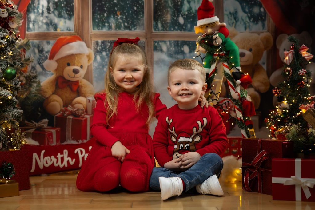 Brother and sister smiling together in matching red Christmas outfits beside a North Pole sign