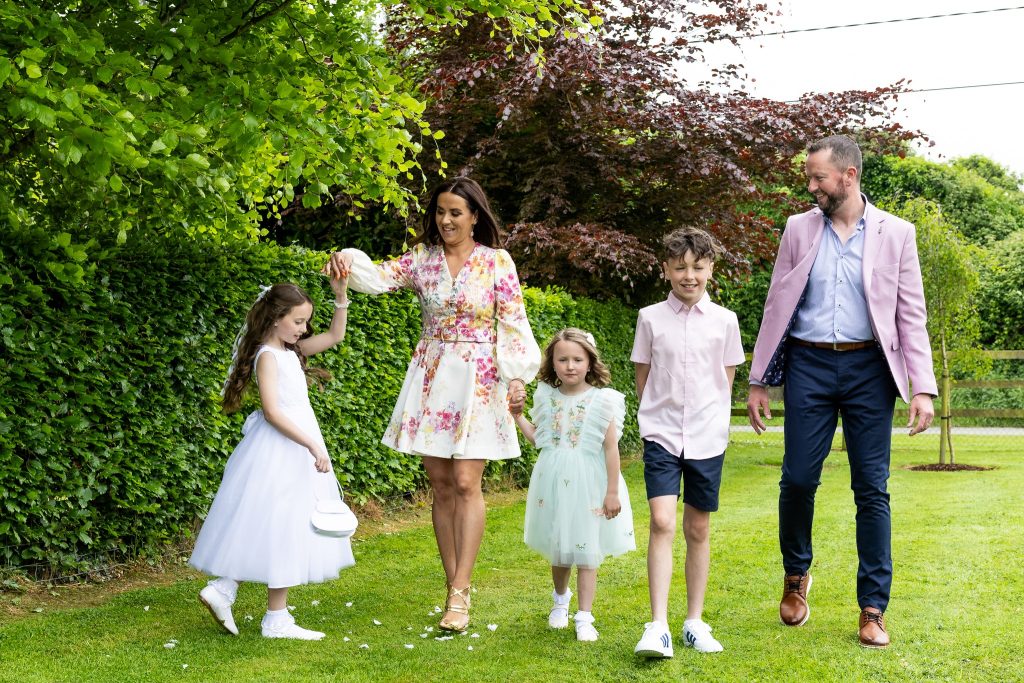 Parents and three children walking together across a garden lawn, the eldest in her white First Communion dress, outdoor family portrait in County Meath